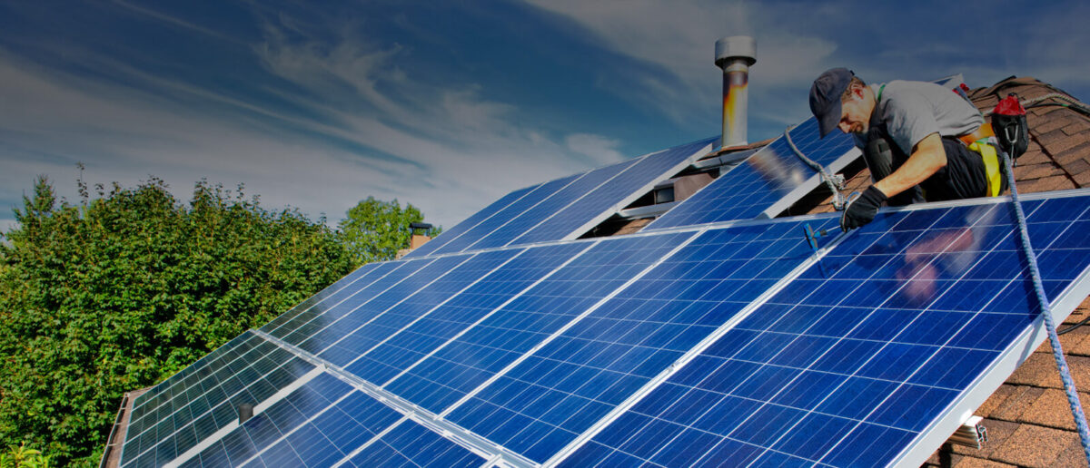 Residential roof with blue solar panels installed, greenery in the background under a clear sky.